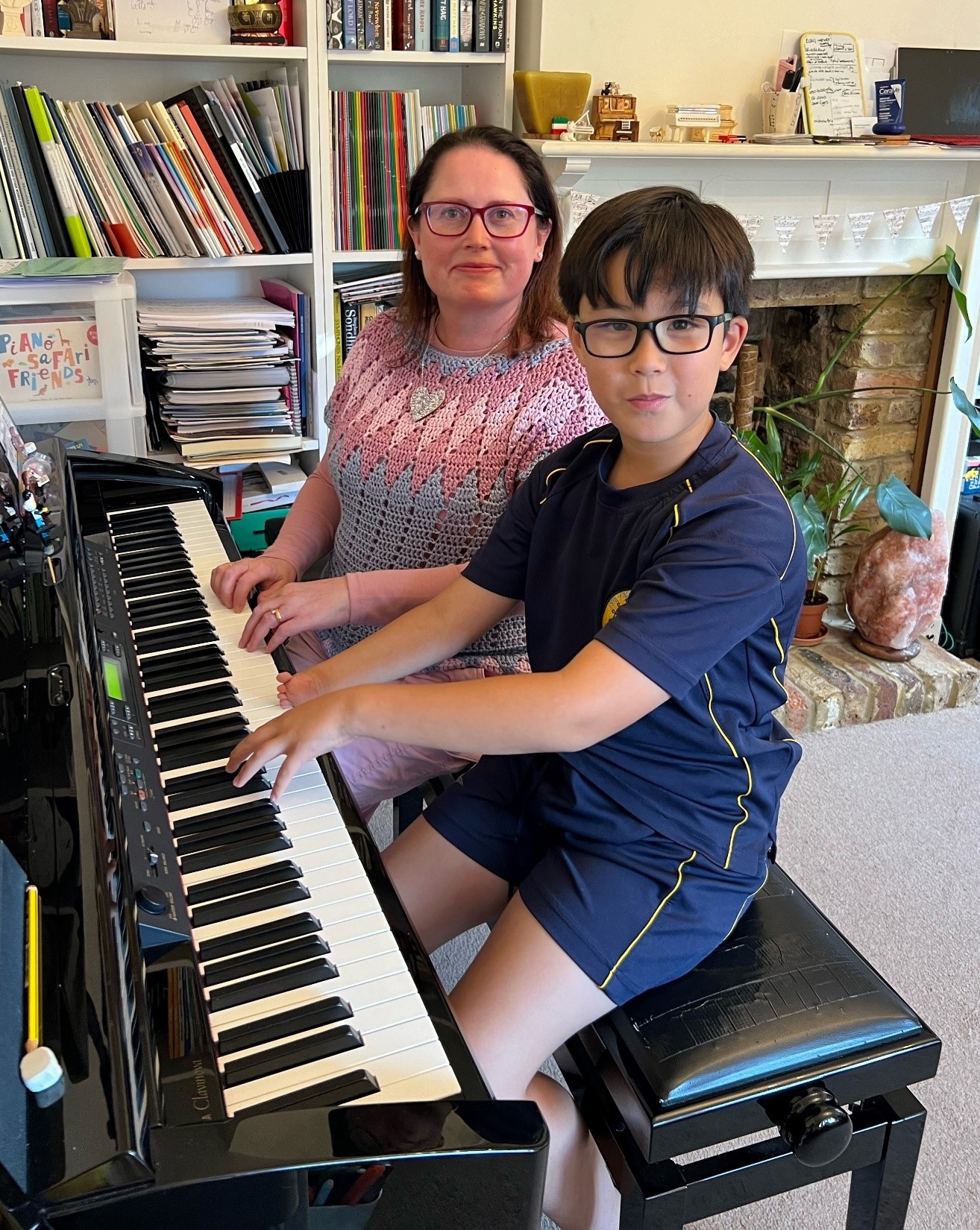 Boy and a woman sitting side by side on at a piano in a living room smiling at the camera. The boy has one hand ready to play the piano keys. His other hand is much smaller and rests on the edge of the keys.