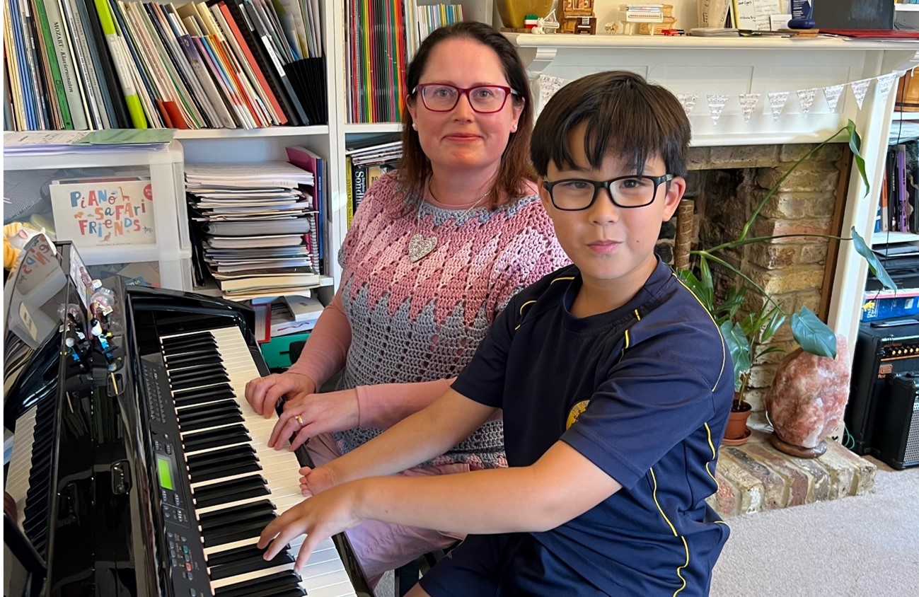 Boy and a woman sitting side by side on at a piano in a living room smiling at the camera. The boy has one hand ready to play the piano keys. His other hand is much smaller and rests on the edge of the keys.