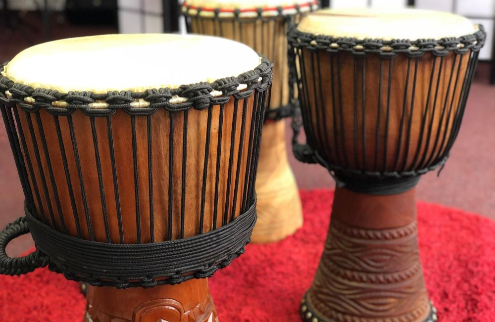 Three drums standing upright on a red carpet. They have light coloured drumskins and dark brown string binding the skins onto the brown wooden bases.